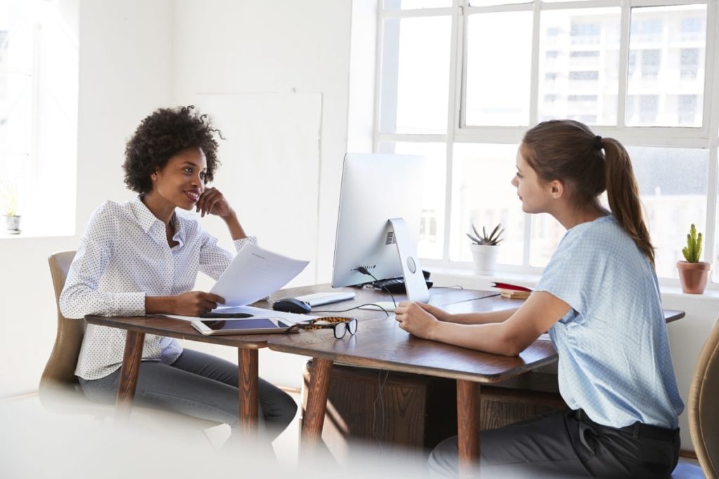 Two young women talking across their desks in an office