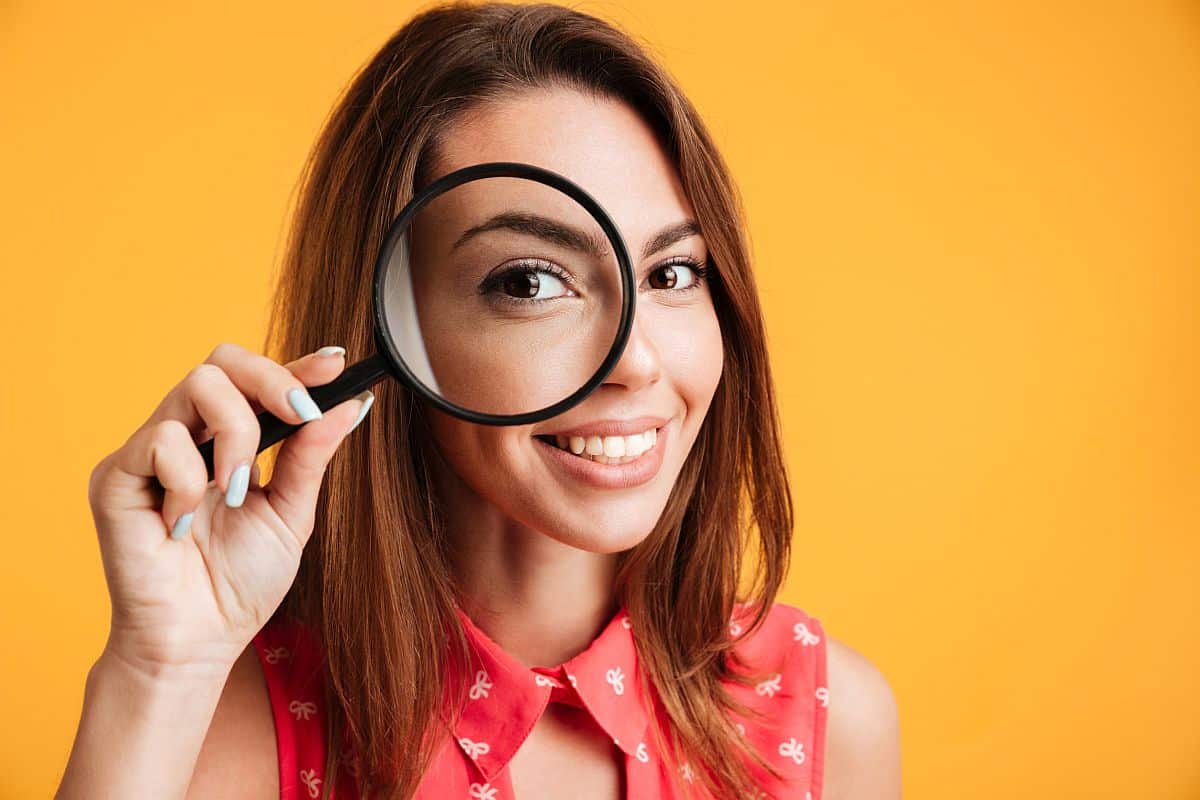 Close up of young cheerful brunette woman looking through magnifying glass; hire for skills and capabilities concept