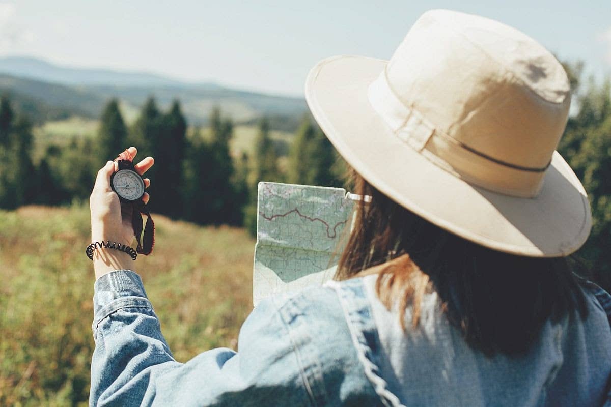 Wanderlust and travel concept. Stylish hipster girl holding map and compass, traveling in sunny mountains. Woman in hat exploring map and hiking on top of mountain on summer vacation.