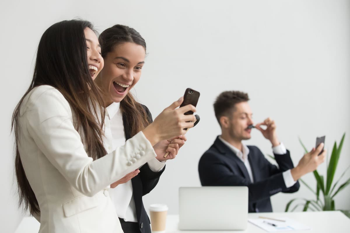 Diverse female colleagues laughing having fun with smartphone; fostering belonging concept