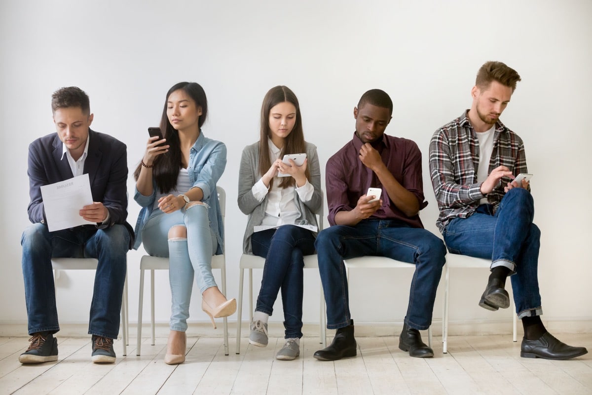 Diverse millennial unemployed people waiting in queue preparing for job interview, multi-ethnic candidates sitting on chairs holding resumes using devices, human resources and employment concept