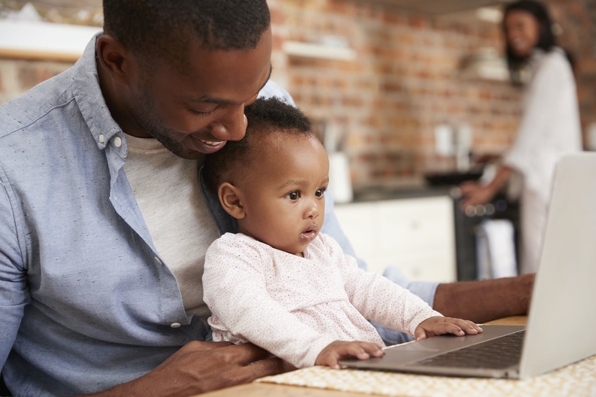 Father And Baby Daughter Use Laptop As Mother Prepares Meal
