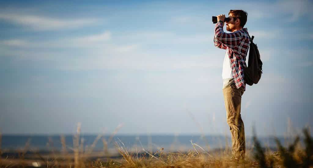 Guy looking at binoculars in hill. man in t-shirt with backpack. Young Caucasian man during hike in valley landscape
