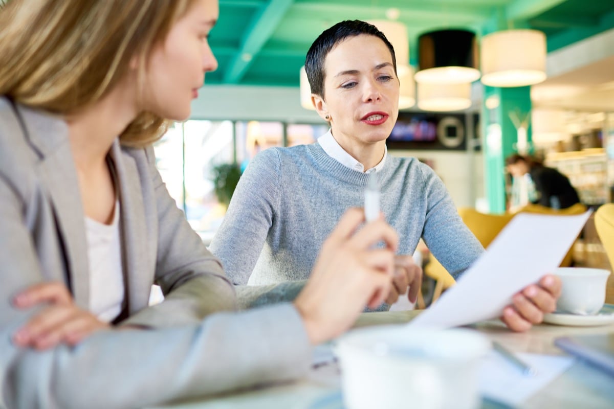 Portrait of two modern businesswomen reading documents sitting at table in cafe and discussing work, focus on serious female boss with short hair